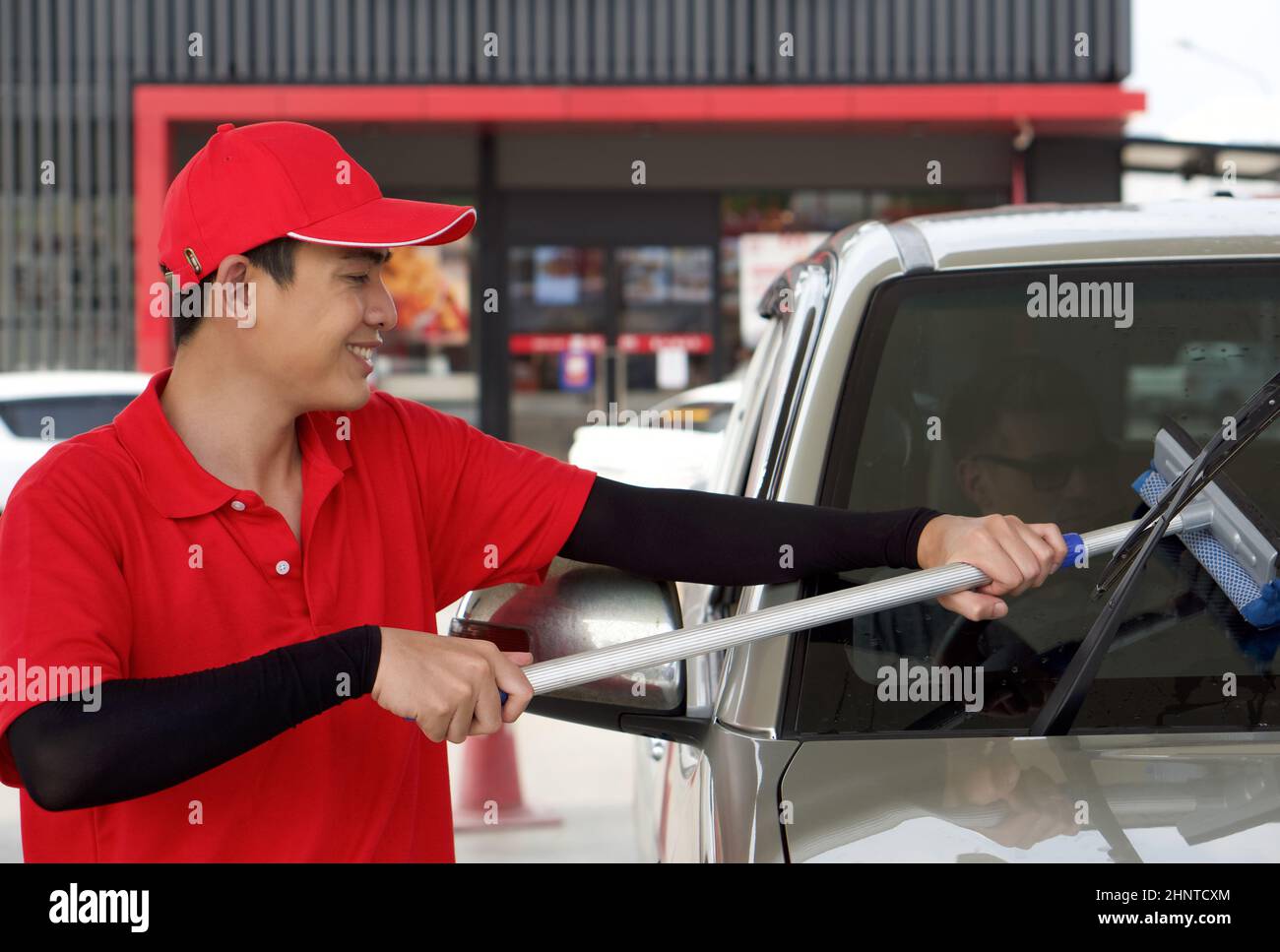Asiatischer Tankstellenmitarbeiter in roter Uniform, der die Windschutzscheibe des Autos mit dem Fensterschieber reinigte. Ein kaukasischer Fahrer mit Sonnenglas wartet im Auto. Stockfoto