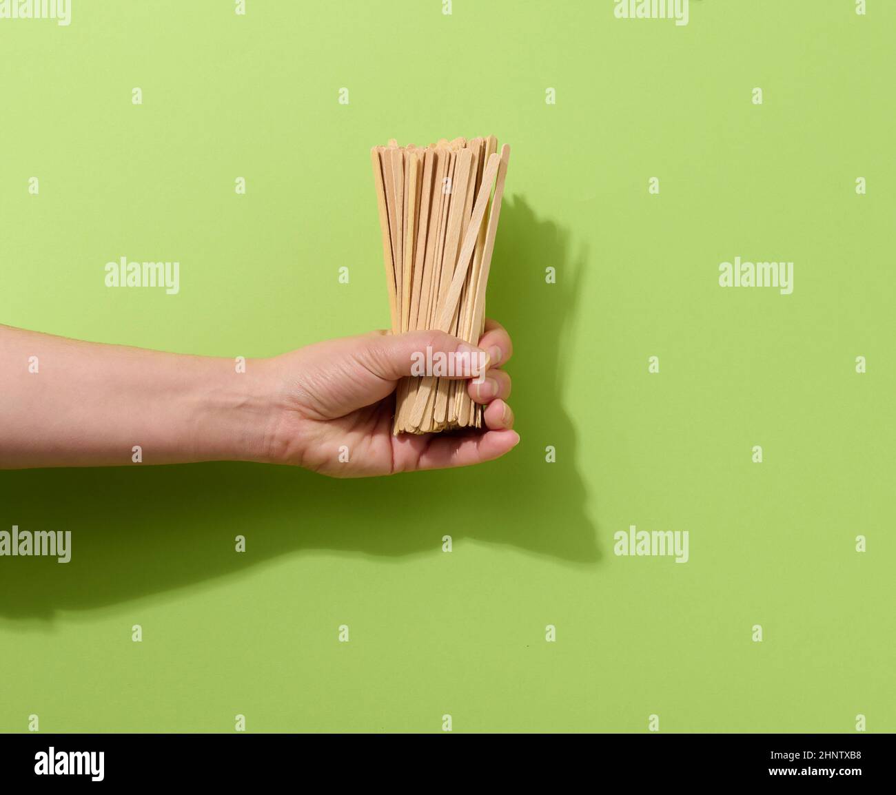 Weibliche Hand hält Einweg-Holzstäbchen zum Rühren von heißen Getränken auf grünem Hintergrund. Kaffee- und Teelöffel, kein Abfall Stockfoto