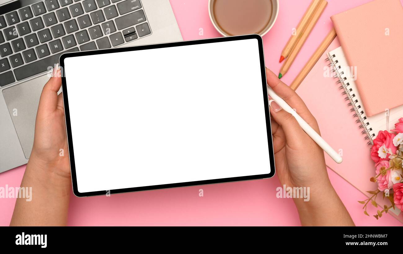 Frau Hände mit digitalen Tablet Touchpad weißen Bildschirm mockup auf rosa pastellfarbenen Hintergrund. Arbeitsplatz mit Laptop, Papeterie, Teetasse und Blumen Bouquet. Stockfoto