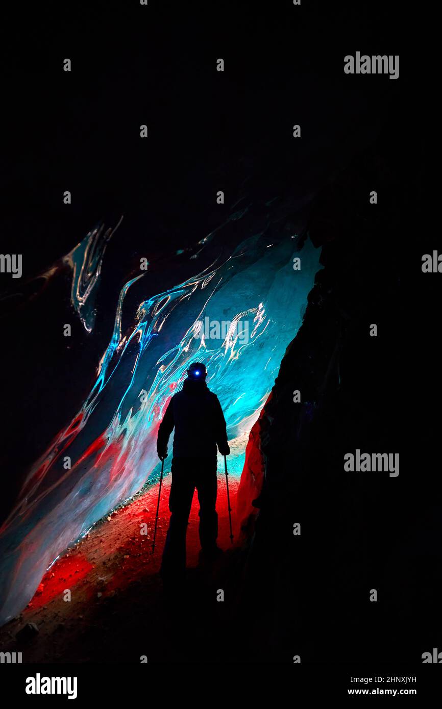 Silhouette von Alpinist in Helm mit rot glühenden Stirnlampe Rückseite in tiefen engen Gletschereishöhle mit cyan Lichter erkunden Winter Berggletscher in Stockfoto