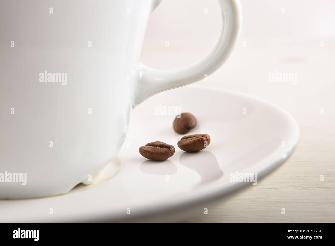Weiße Porzellanbecher mit Kaffee auf weißem Teller mit Bohnen auf weißem Holztisch beim Frühstück Makroansicht. Vorderansicht. Horizontale Zusammensetzung. Stockfoto