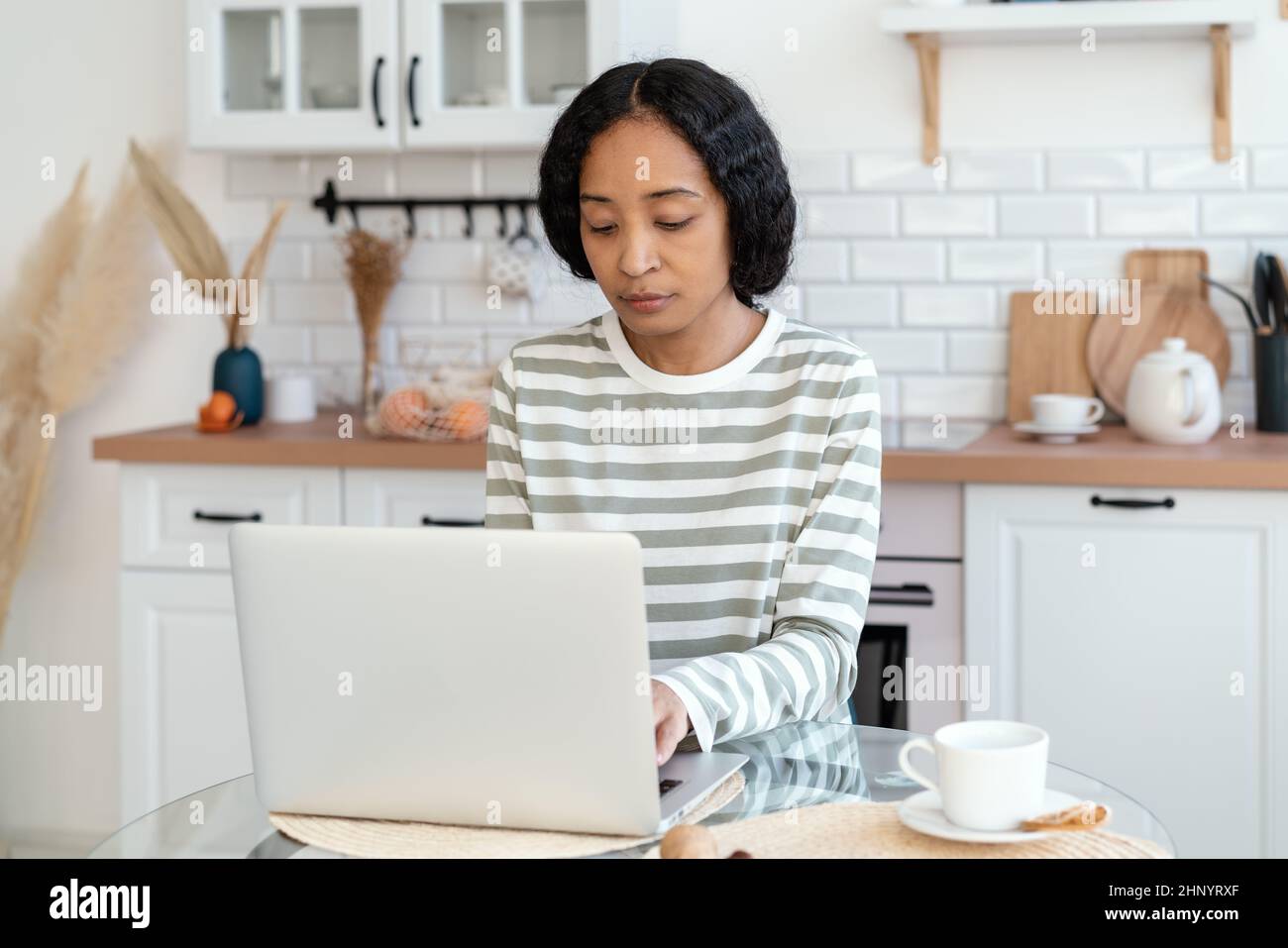afroamerikanische Frau, die in der Küche sitzt und einen Laptop benutzt. Selbstbewusst und konzentriert auf Remote-Arbeit Stockfoto