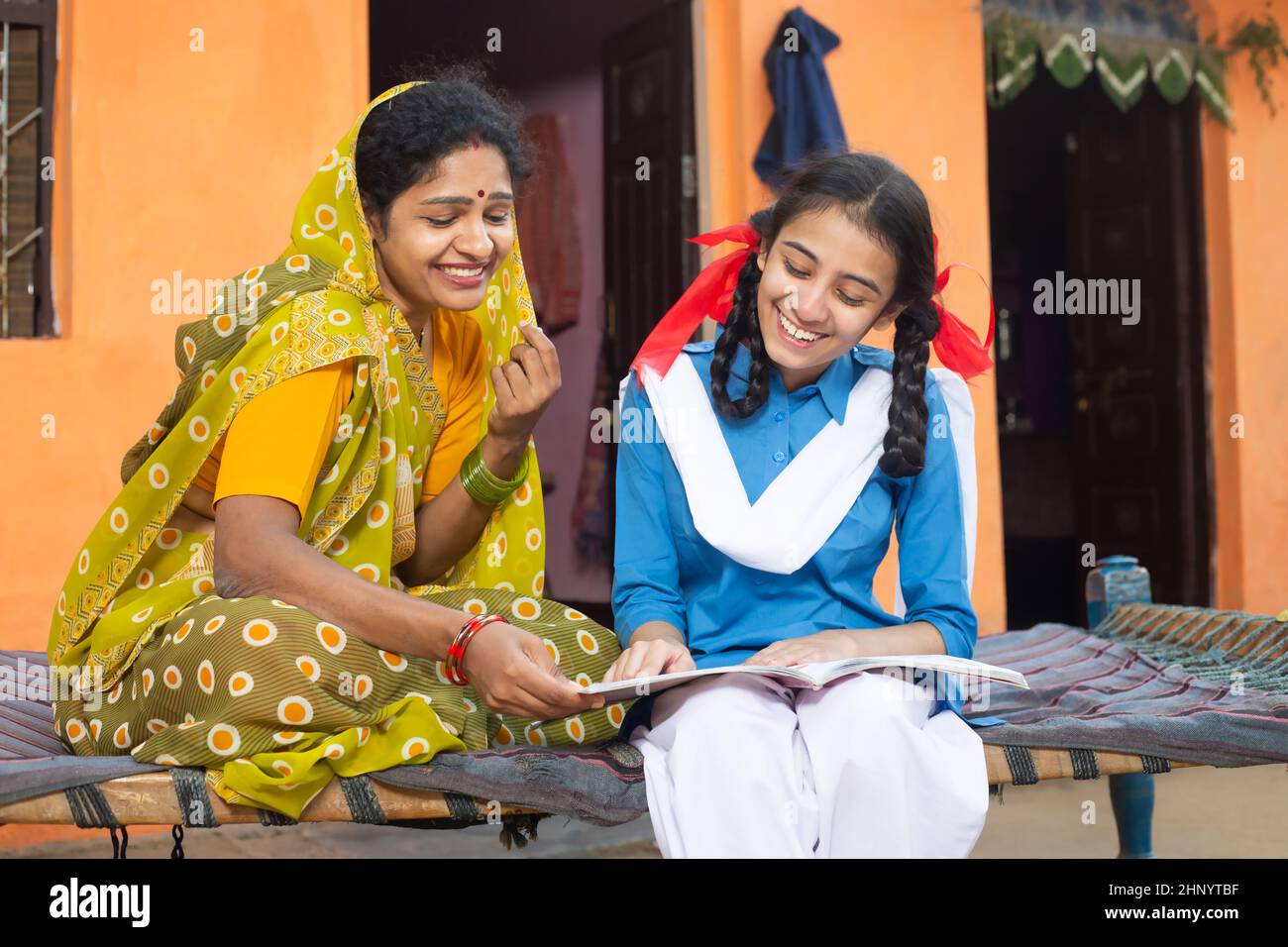 Glückliche traditionelle indische Mutter hilft ihrer jungen Tochter in Studien, ländliche Mädchen Kind hält Notebook zu Hause studieren. Frau trägt Sari Teachin Stockfoto