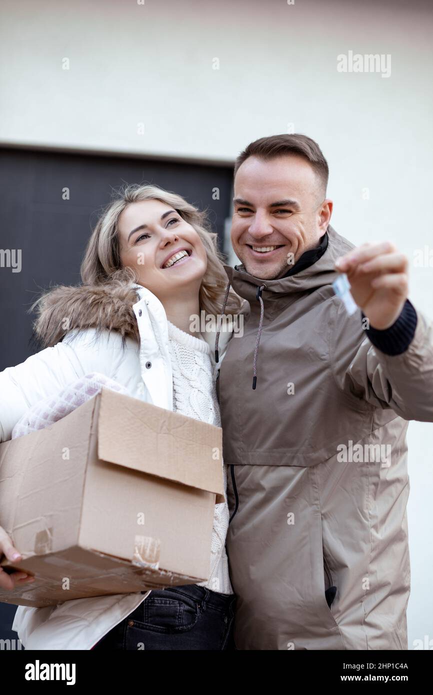 Porträt eines lächelnden Paares in legeren Oberkleidern stehen umarmt, halten Schlüssel und Pappkarton vor dem Haus. Stockfoto