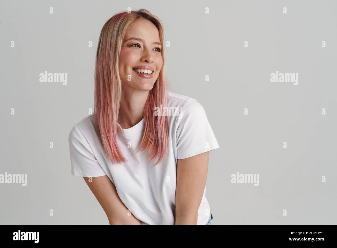 Glücklich lächelnde junge blonde weiße Frau in legerer Kleidung, die über grauem Wandhintergrund steht und zur Seite schaut Stockfoto