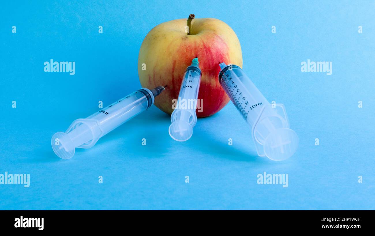 Drei verschiedene medizinische Spritzen, die tief in einen Apfel auf blauem Hintergrund stecken. Die Frucht ist mit Spritzen übersät. Ein reifer roter Apfel, der von al durchbohrt wurde Stockfoto