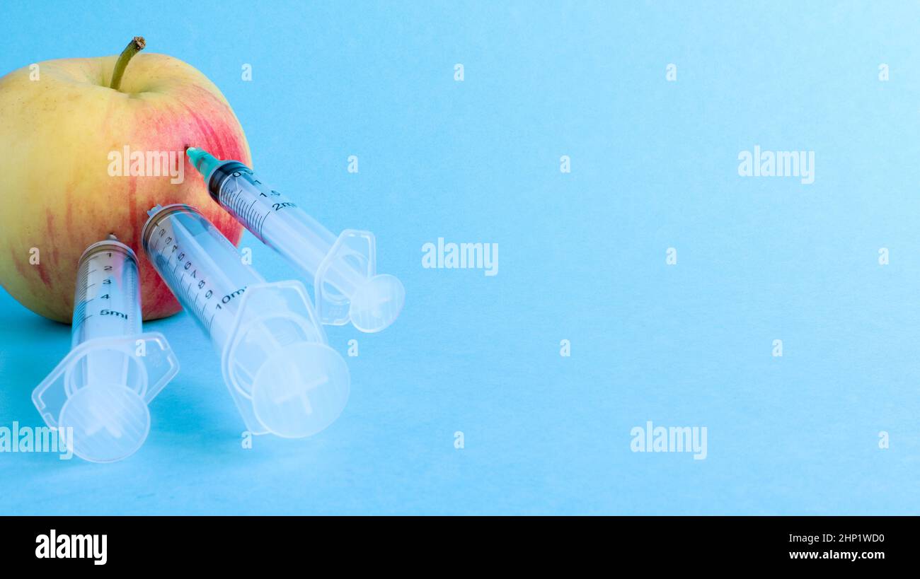 Drei verschiedene medizinische Spritzen, die tief in einen Apfel auf blauem Hintergrund stecken. Die Frucht ist mit Spritzen übersät. Ein reifer roter Apfel, der von al durchbohrt wurde Stockfoto