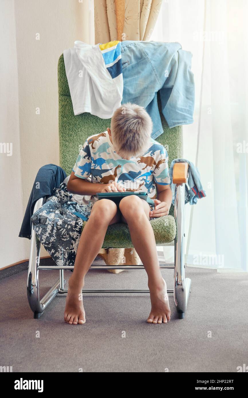 Blonde junge sitzt in Sessel mit Haufen von schmutzigen Kleidern und spielt Online-Spiele über Tablet. Schüler verbringt Freizeit mit modernen Technologien Stockfoto