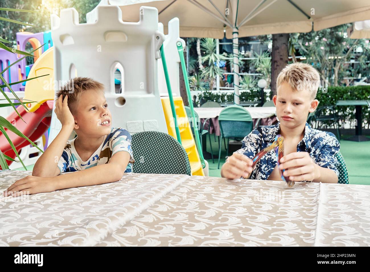 Blonde Brüder sitzen am Restauranttisch und spielen auf das Mittagessen. Schüler sitzen in Langeweile gegen Unterhaltungszone mit Plastikfestung und rutschte Stockfoto