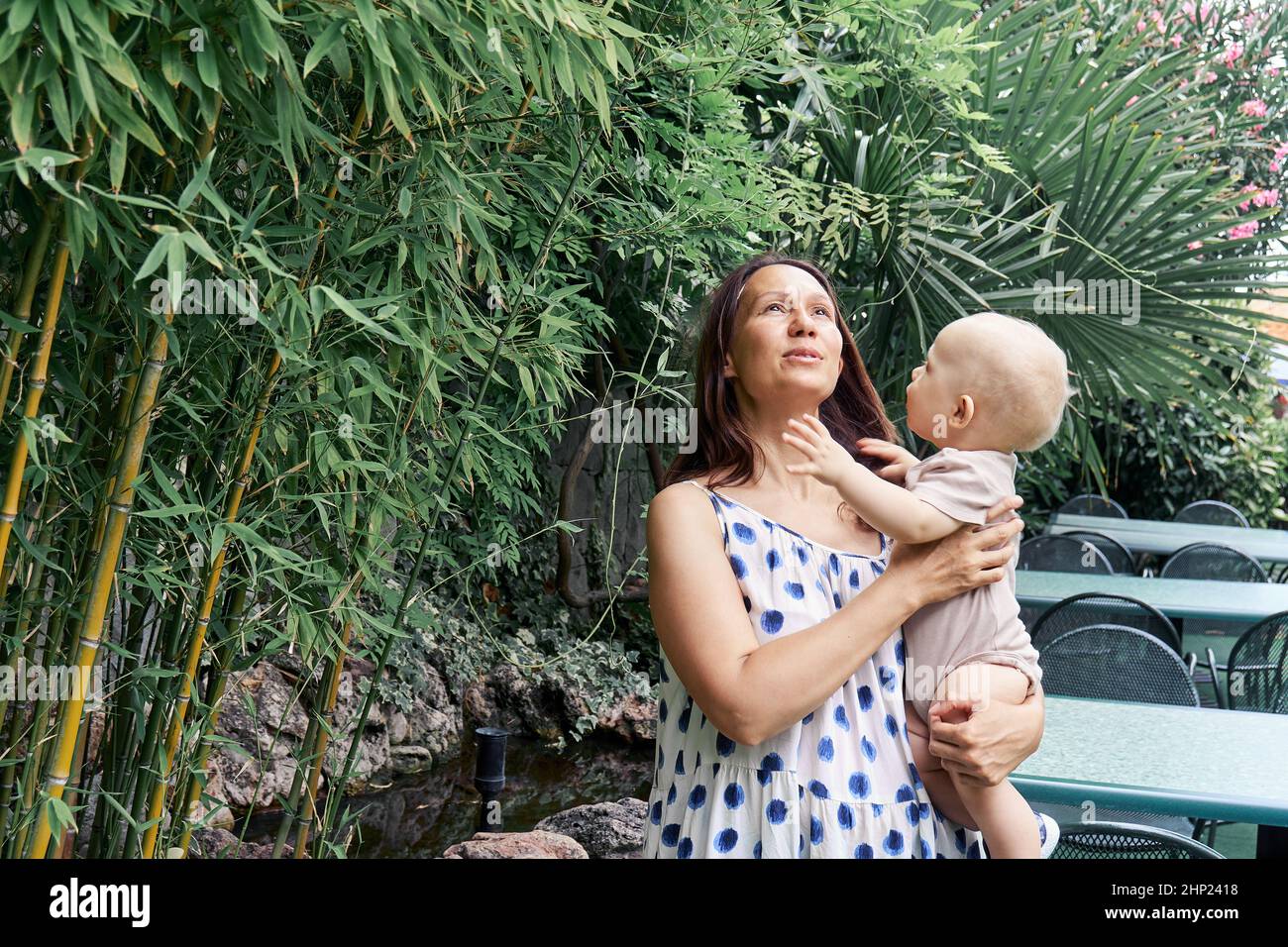 Die junge Mutter hält ein blondes Baby in Armen, das im Bambuswald spazieren geht. Dunkelhaarige Frau und Kind genießen frische Luft und entspannen sich im Urlaub in den Tropen Stockfoto
