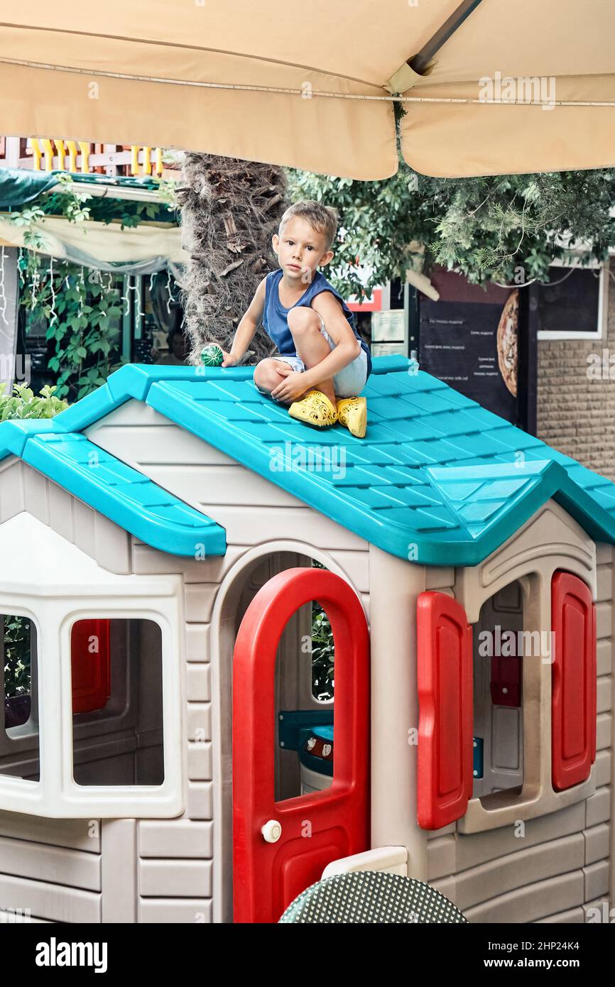 Vorschulkinder in Sommerkleidung sitzen auf dem hellen Dach des Spielzeughauses im Spielbereich des Restaurants. Das blonde Kind verbringt seine Freizeit damit, draußen zu spielen Stockfoto
