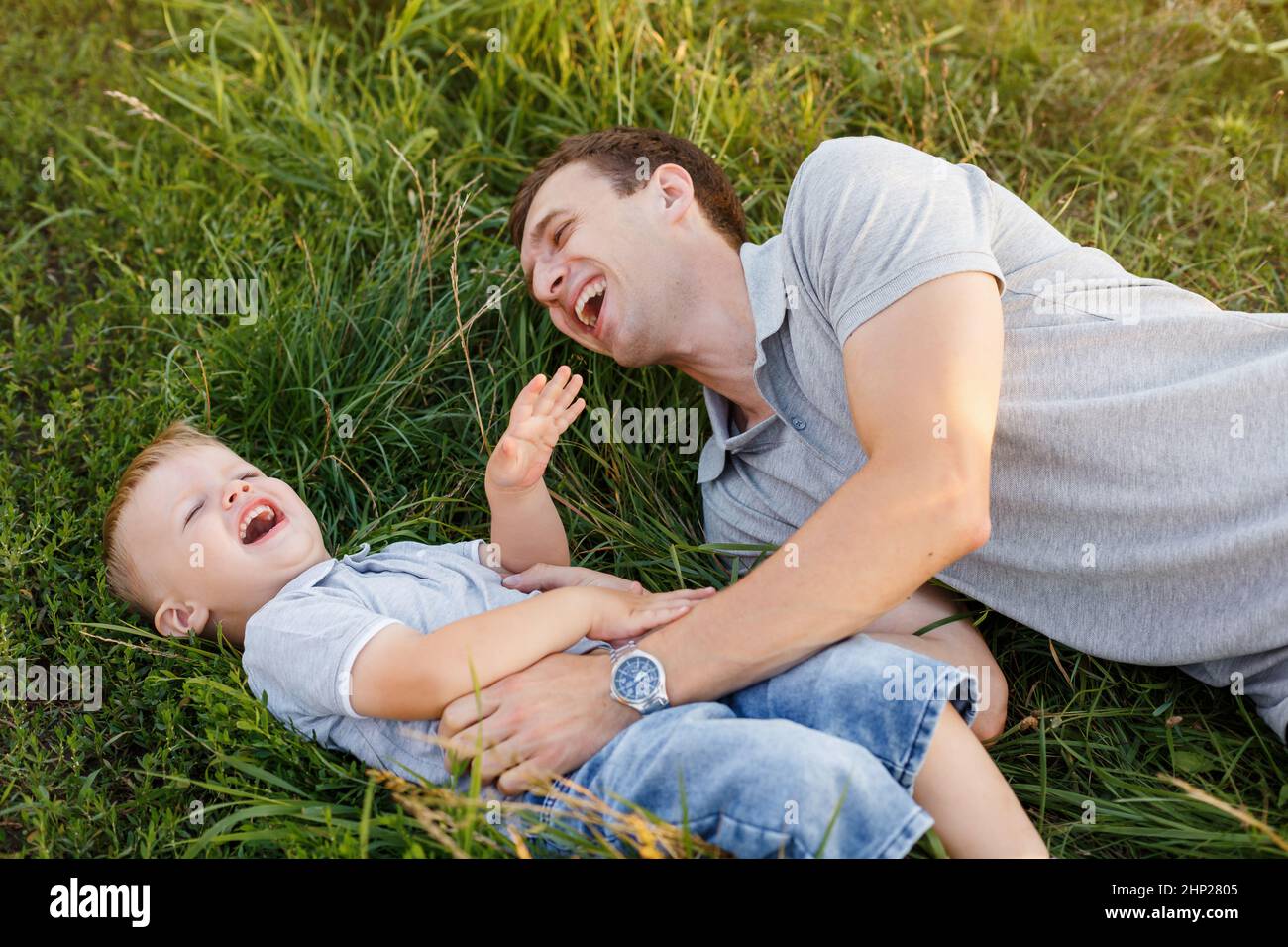 Vater und Sohn kitzeln sich gegenseitig auf dem Gras. Kleiner Junge und sein Vater haben Spaß und lachen. Sommer im Freien Lifestyle, Familie Freizeit, Elternschaft Stockfoto