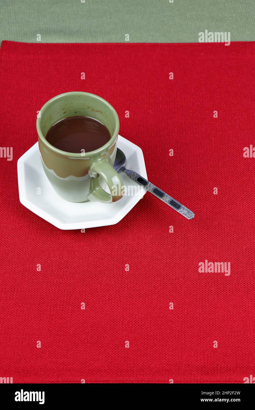 Eine Portion heißer Schokolade-Gourmet-Mischung in einem Kaffeebecher, der auf einer weißen achteckigen Untertasse auf einer roten Platzmatte mit einem grünen Tischtuch dahinter ruht. Liqu Stockfoto
