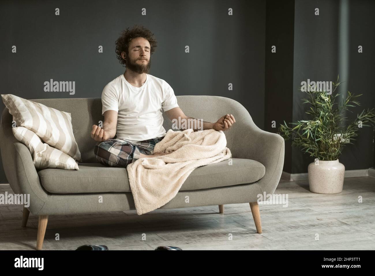 Junger, gutaussehender Mann, der zu Hause auf der Couch oder dem Sofa sitzt und Meditation, Entspannung und Ruhe genießt, in seinem Schlafanzug im Wohnzimmer während des Lockdowns oder des Krankseins. Stockfoto