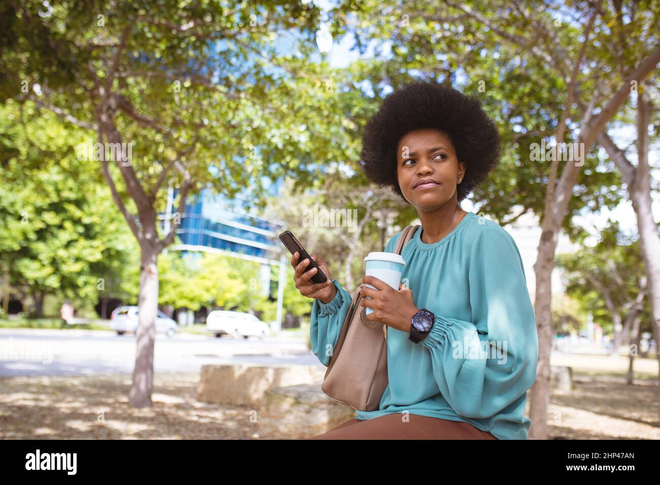 afroamerikanische Geschäftsfrau mit mittlerem Erwachsenenalter schaut weg, während sie mit einem Telefon und einer Einweg-Tasse sitzt Stockfoto
