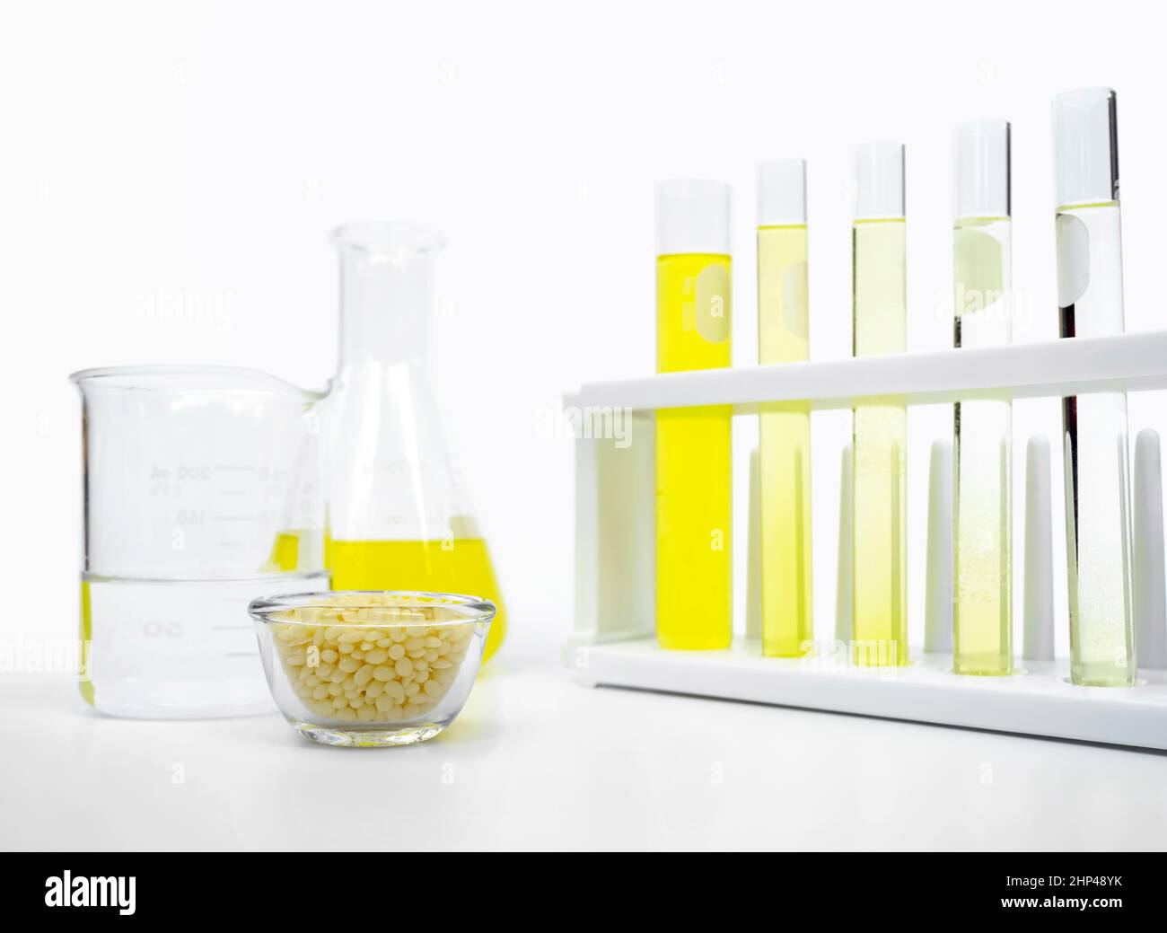Candelilla-Wachs im Glasbehälter und gelbe kosmetische Farbe (Öl) im erlenmeyerkolben neben Reagenzglasständer und Becher platzieren. Stockfoto
