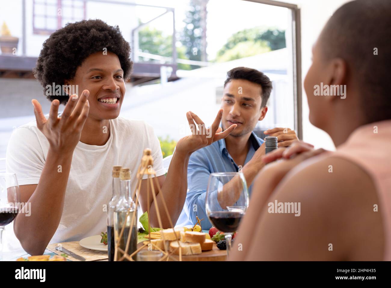 Glücklicher junger Birazialmann mit afro-Frisur, der mit Freunden spricht, die zusammen am Esstisch sitzen Stockfoto