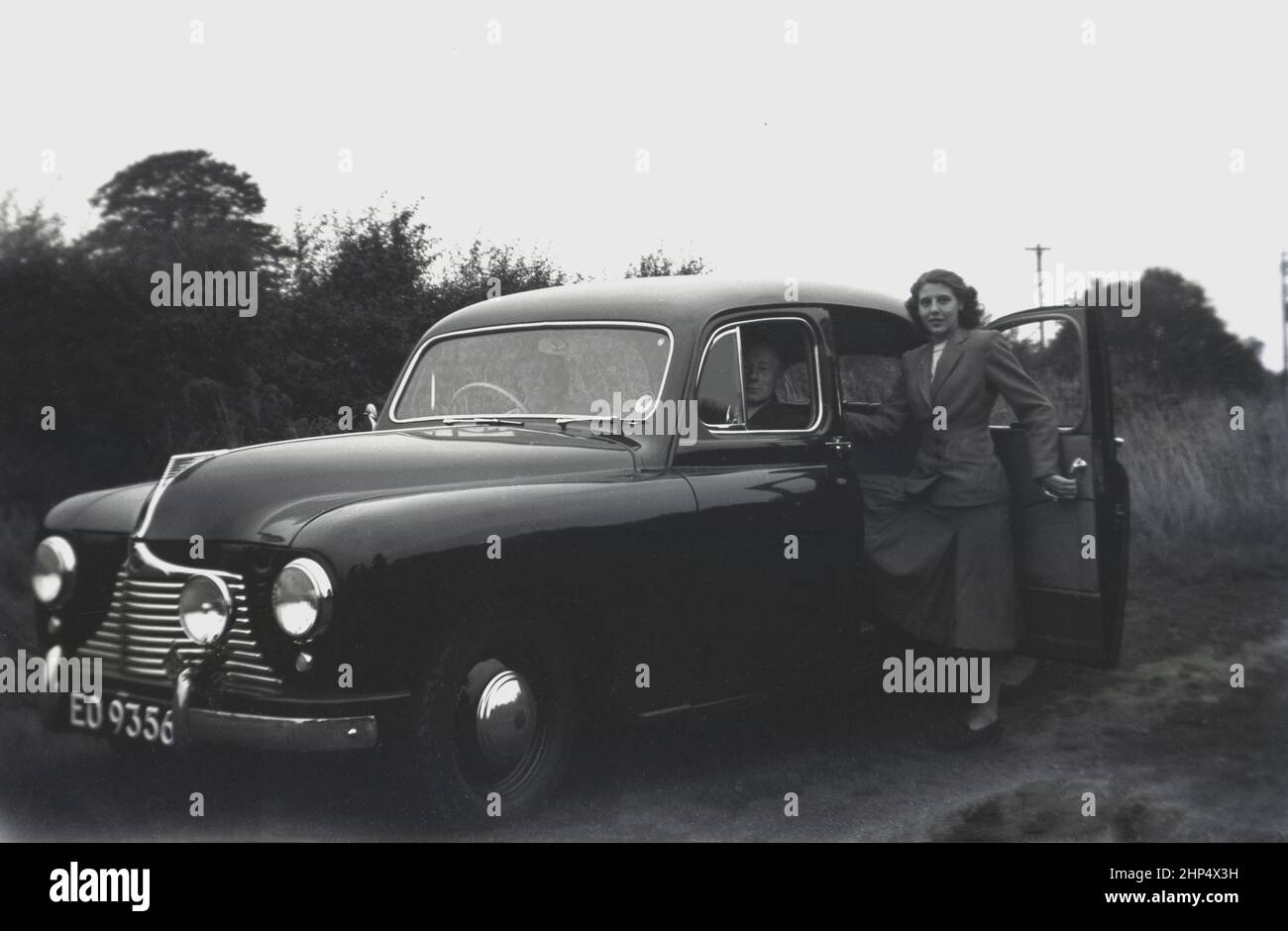 1940s, historisch, draußen auf einer Counrryside-Strecke, eine Dame, die kurz davor steht, auf den Rücksitz eines neuen Autos zu steigen, eines Singer SM1500, England, Großbritannien. Die Hintertüren öffnen sich aus der Mitte, was als "Suizidtüren" bekannt wurde, da man leicht aus ihnen herausfallen könnte. Die SM1500 ist eine kleine Familienlimousine mit 4 Türen, die zwischen 1948 und 1956 produziert wurde und von Singer Motors, einem britischen Automobilunternehmen, das 1874 in Coventry, England, von George Singer gegründet wurde, hergestellt wurde. Ursprünglich ein Hersteller von Fahrrädern, begann das Unternehmen 1901 mit der Herstellung von Motorwagen. Stockfoto