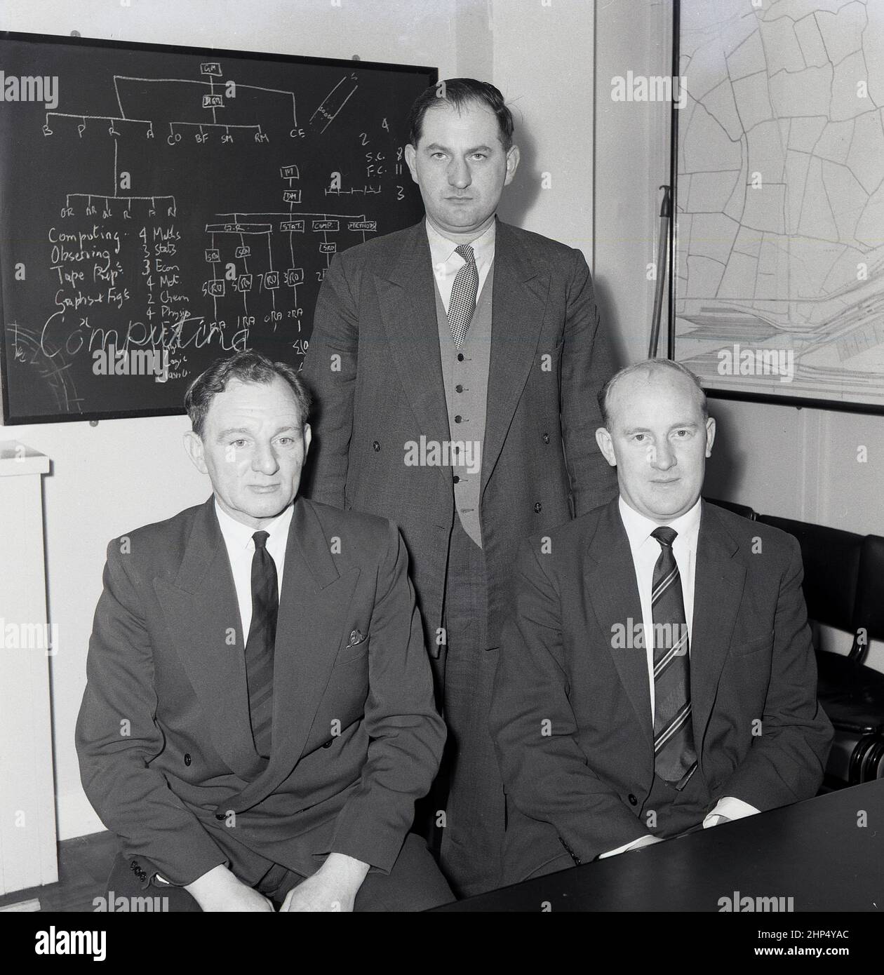 1960s, historische, zwei Leiter der Stahlfabrik und ein Ausbilder, die für ihr Bild mit einer kleinen Tafel hinter ihnen sitzen und Details ihrer Lektion enthalten. Das Wort Computing wird über die Tafel geschrieben. Die Stahlindustrie war ein früher Anwender von Computern für die Analyse von Metallen, die Prozesssteuerung von Öfen und die Auftragsabwicklung. Der erste Leo II-Computer, der außerhalb des britischen Tee- und Kaffeeunternehmens Lyons installiert wurde, war 1958 in einem Stahlwerk in Corby, England, Großbritannien. Stockfoto