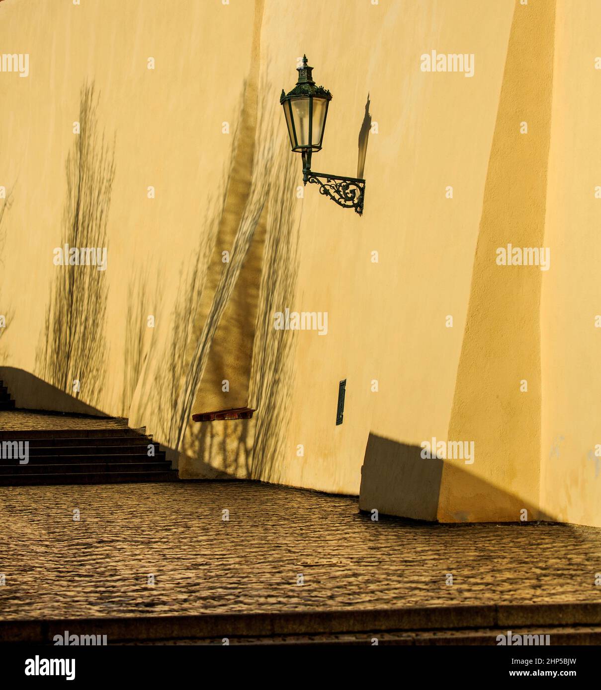 Ein gepflasterter Durchgang mit der gelben Pfeilermauer der Prager Burg und einer traditionellen verzierten Straßenlaterne, an einem sonnigen Tag mit Schatten, Prag, Tschechien Stockfoto