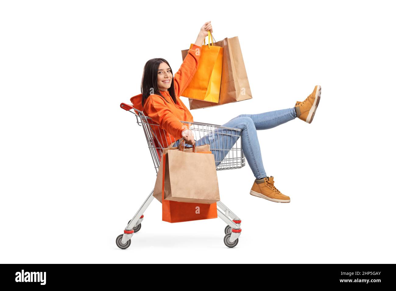 In voller Länge Porträt einer glücklichen jungen Frau, die in einem Einkaufswagen sitzt und Einkaufstaschen auf weißem Hintergrund isoliert hält Stockfoto