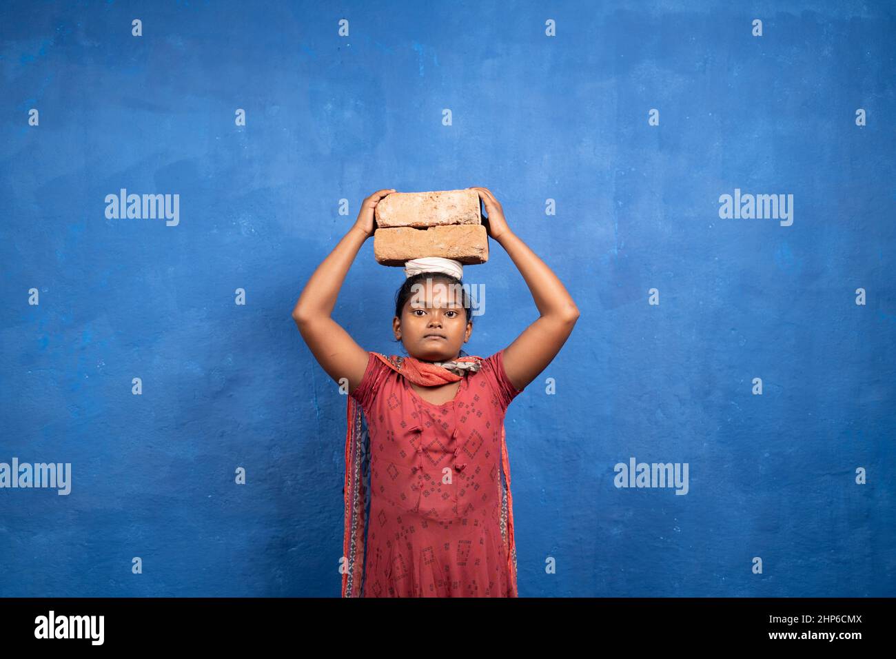 Junges Mädchen, das Steine auf dem Kopf hält, indem es die Kamera mit dem Kopierraum betrachtet - Konzept von Kinderarbeit, Armut und Unwissenheit. Stockfoto