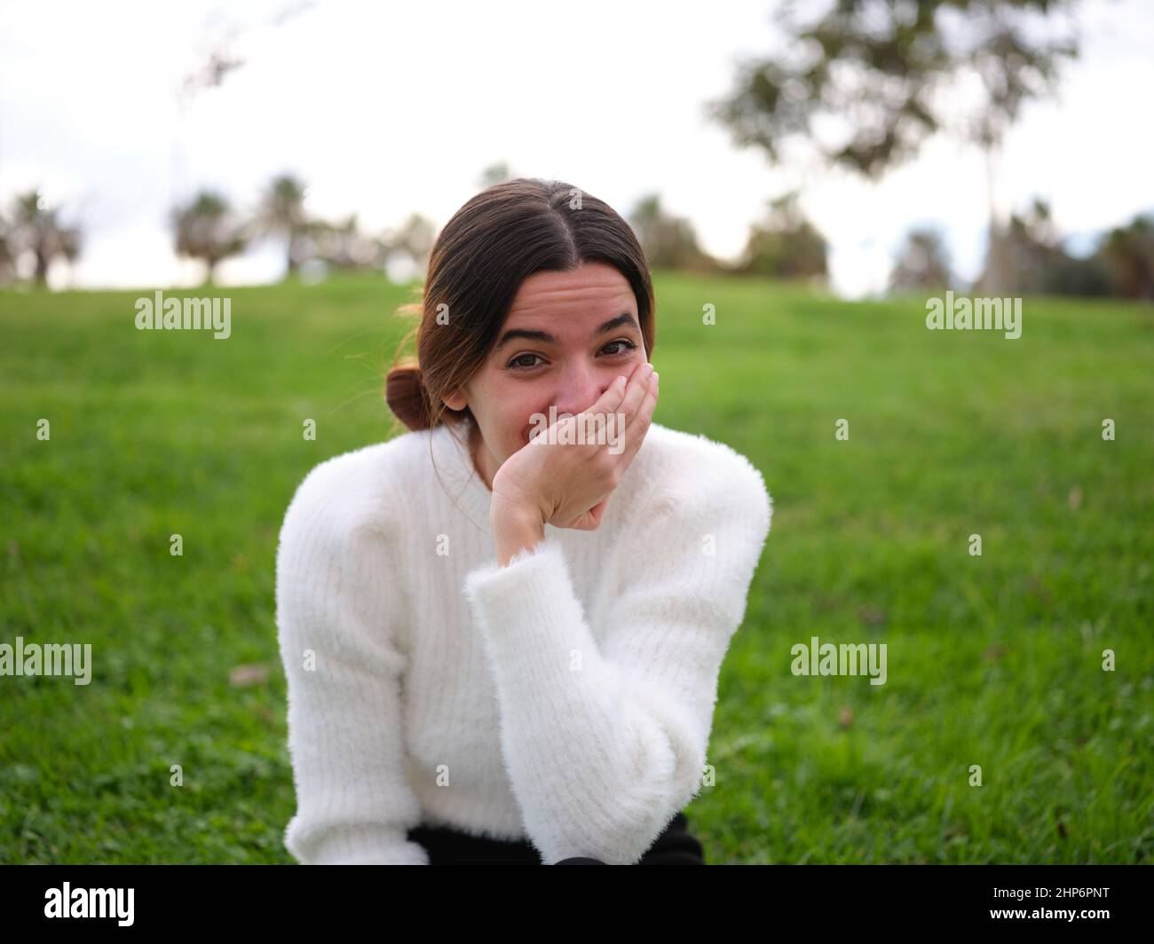 Eine fröhliche junge Frau im Park, die lachend auf die Kamera schaut, die ihren Mund mit der Hand bedeckt. Stockfoto
