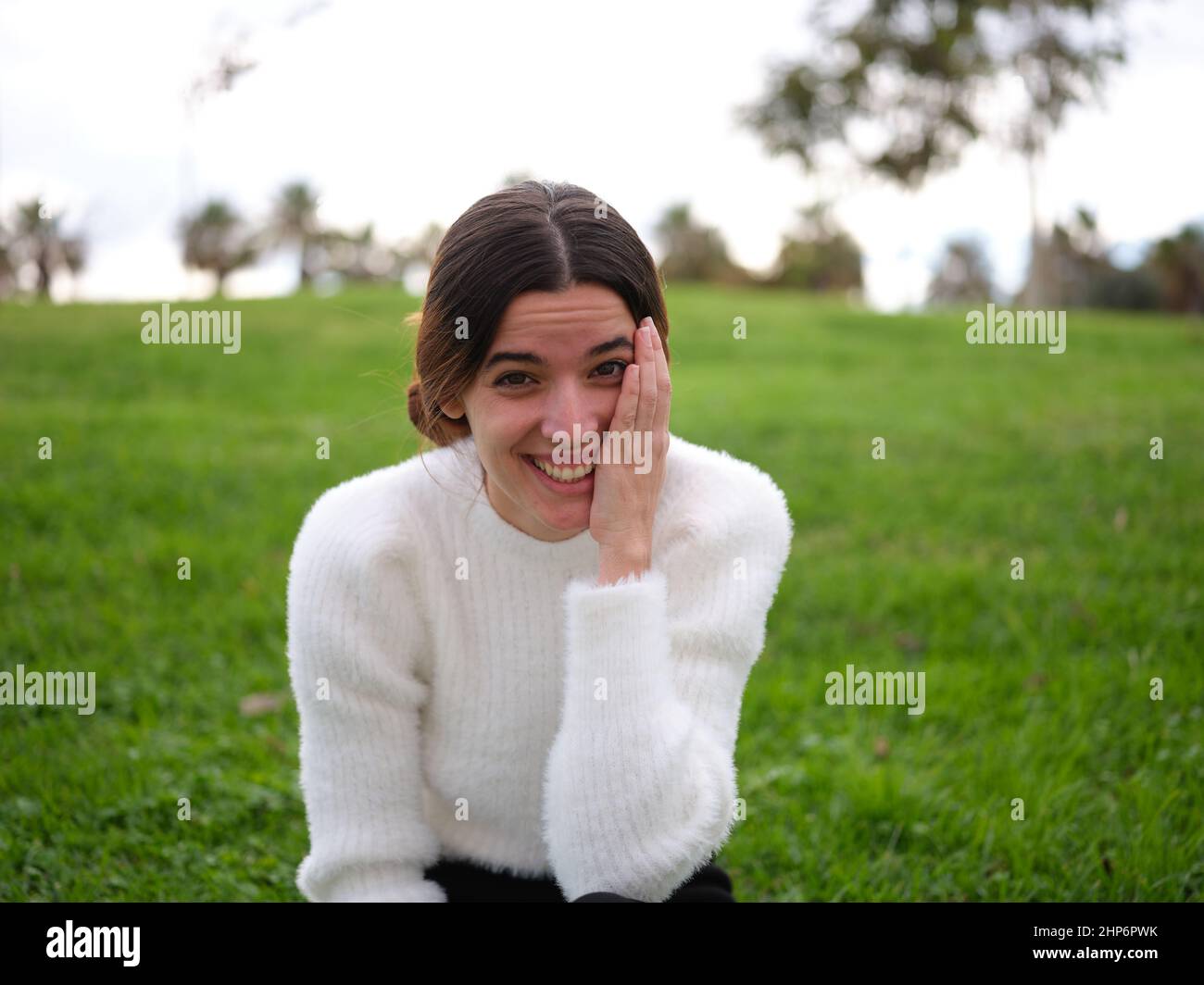 Eine glückliche junge Frau im Park, die die Kamera anschaut und ihr Gesicht mit der Hand berührt, während sie lacht. Stockfoto