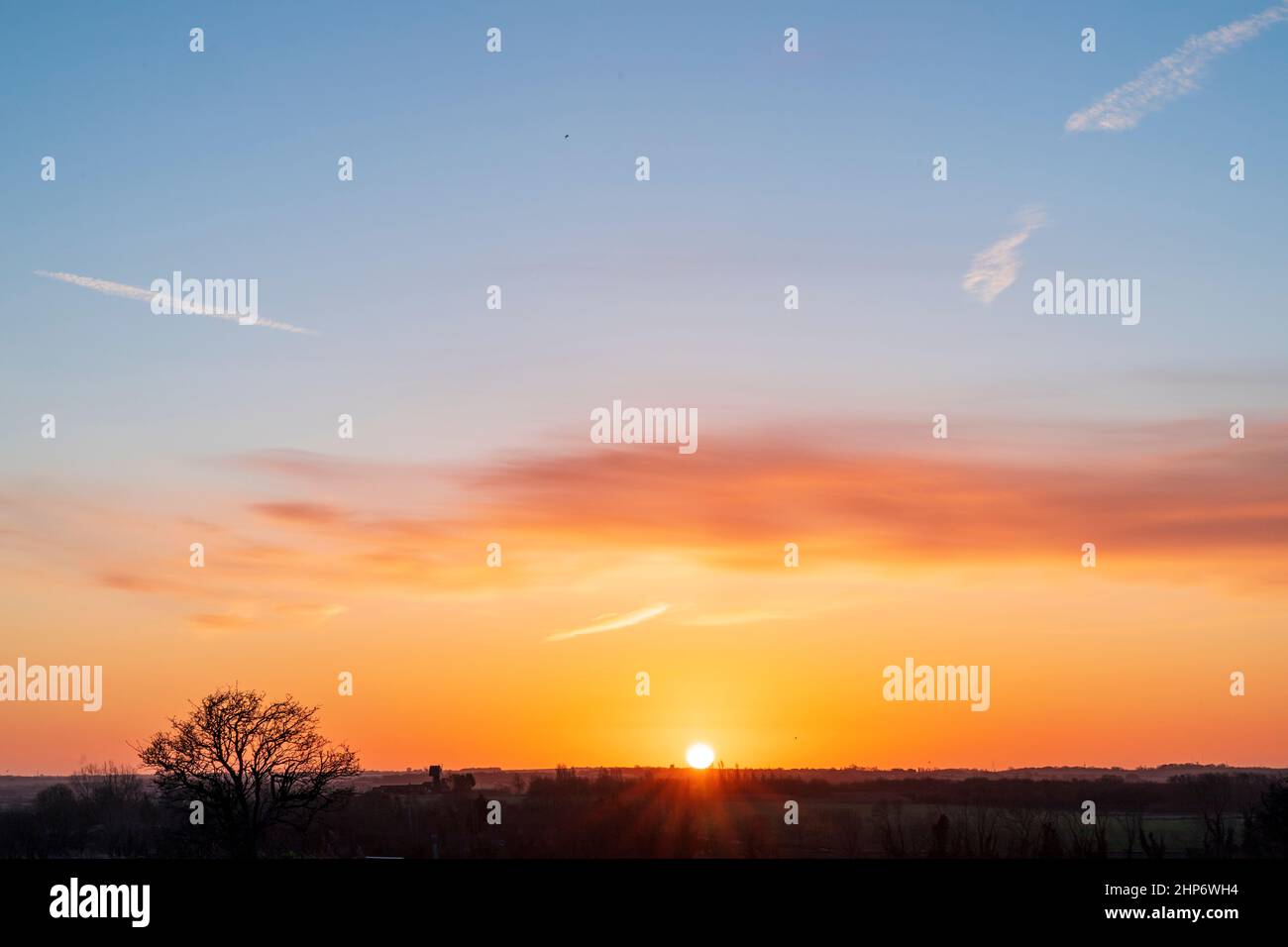 Sonnenaufgang über einer ziemlich flachen Landschaft mit ein paar verstreuten Bäumen in Kent im Winter. Blick von Hillborough in Richtung Ramsgate zum Sandwich-Gebiet. Die Sonne steigt in einen orangefarbenen Himmel auf und wird höher zu blau mit einigen verstreuten dünnen Wolken. Stockfoto