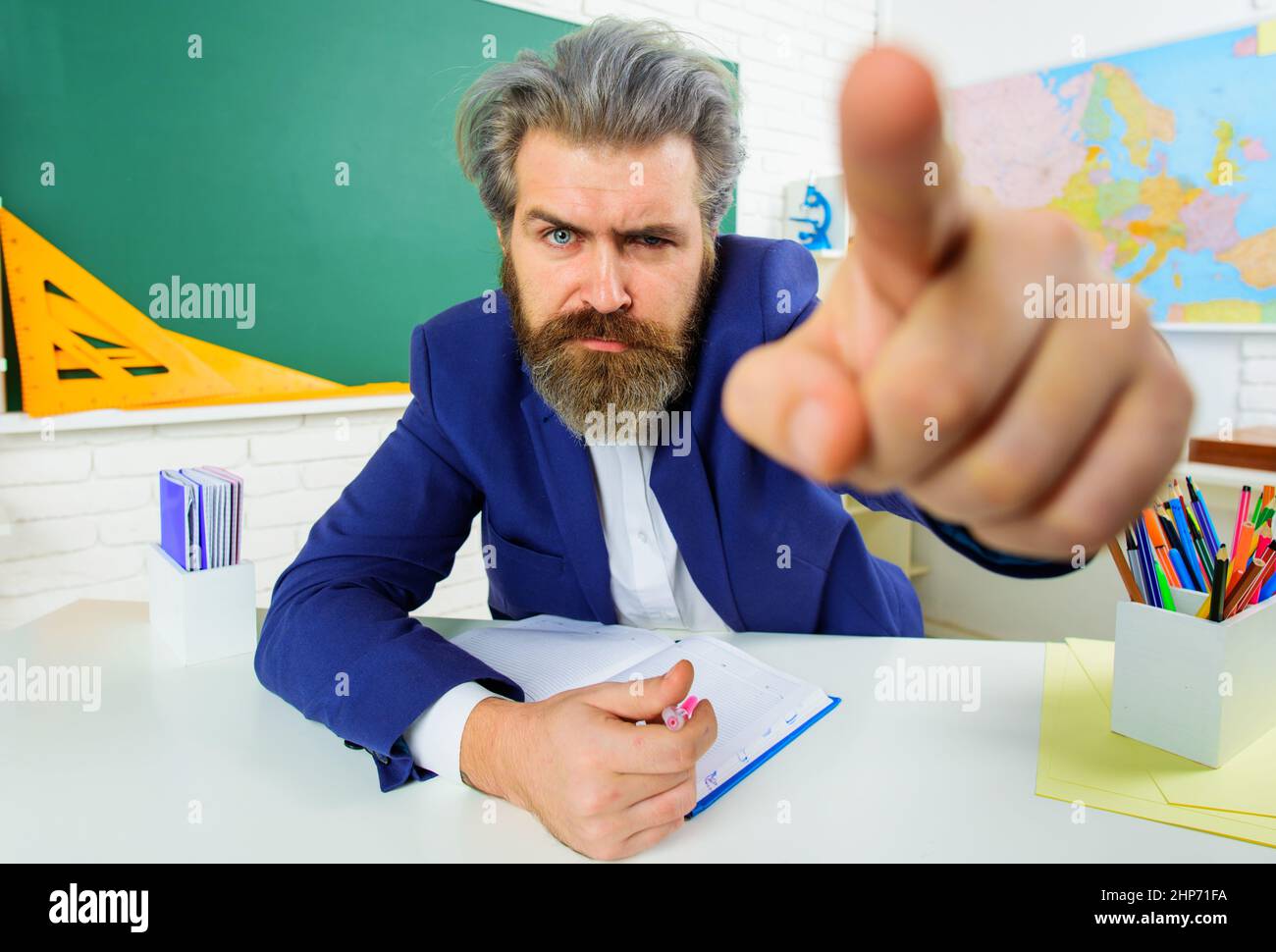 Ernsthafter Lehrer im Klassenzimmer zeigt den Finger. Bärtiger Mann, der in der Klasse am Tisch sitzt. Lernen, Bildung. Stockfoto