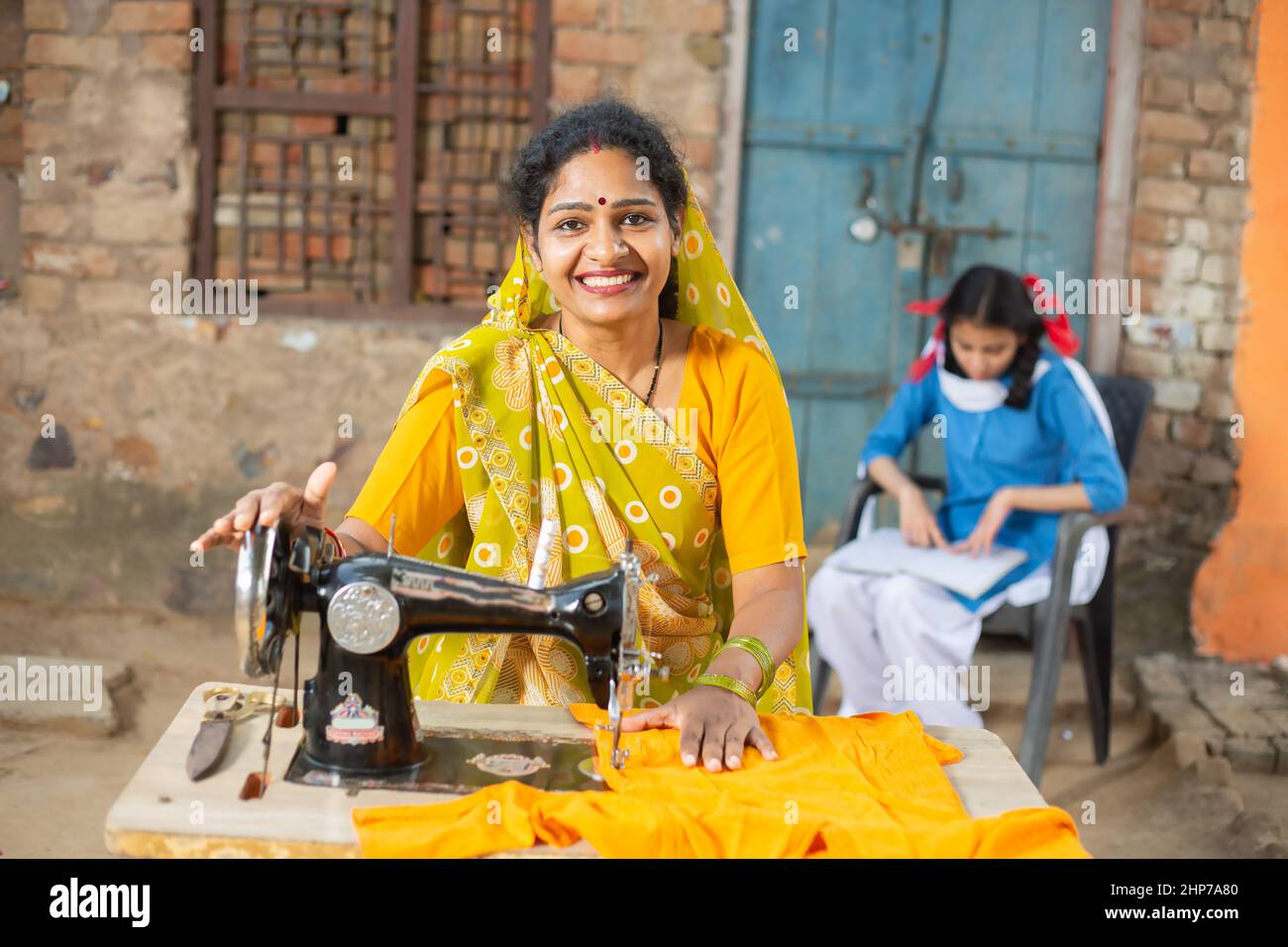 Porträt einer glücklichen, traditionellen indischen Frau, die mit einer Nähmaschine einen Sari trägt, während ihre junge Tochter hinter ihr in Schuluniform studiert. Stockfoto