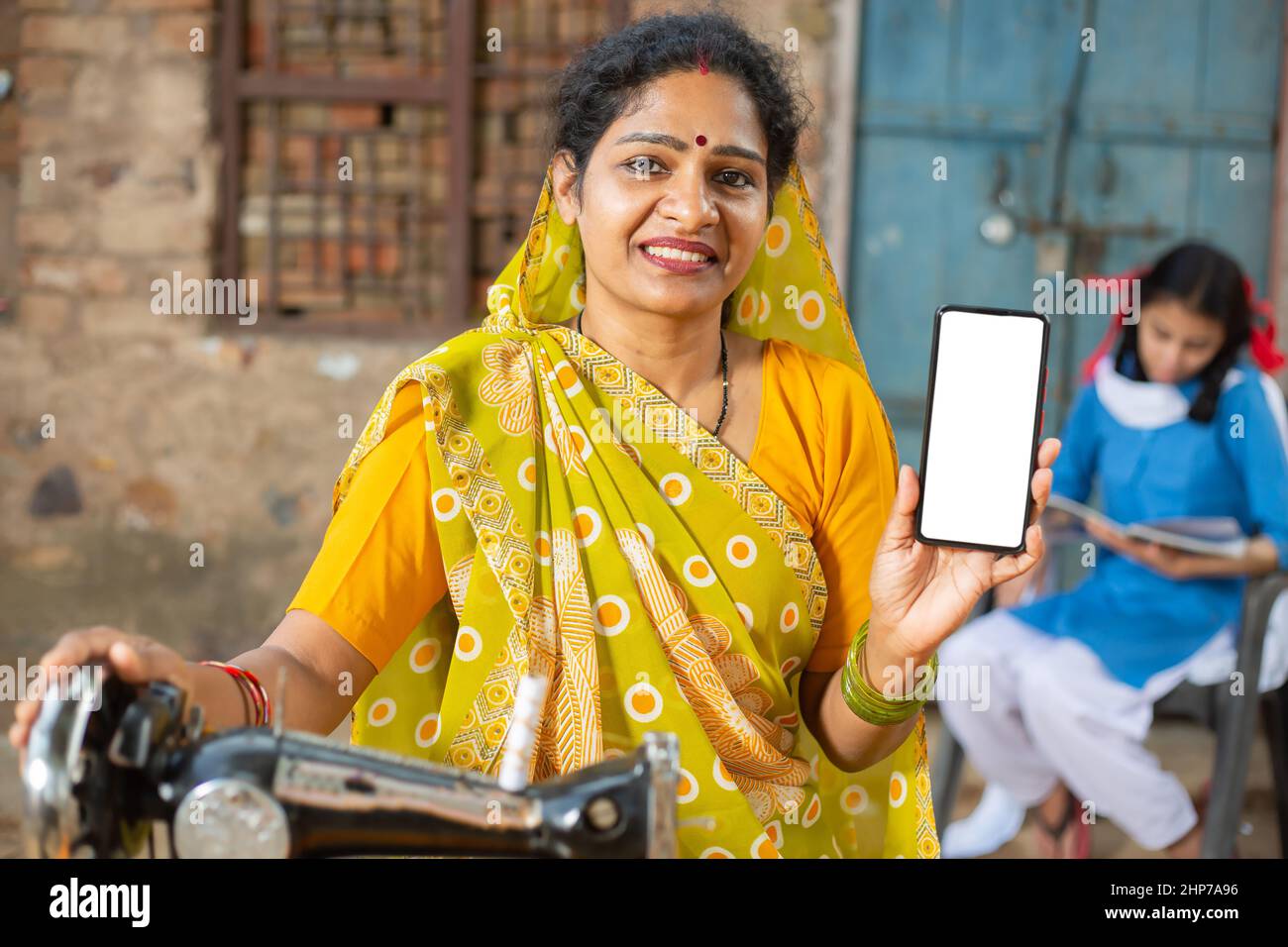 Porträt einer glücklichen traditionellen indischen Frau, die mit einer Nähmaschine einen Sari trägt, während sie ein Smartphone mit leerem Display zeigt, um Werbung zu schalten, weiblich Stockfoto