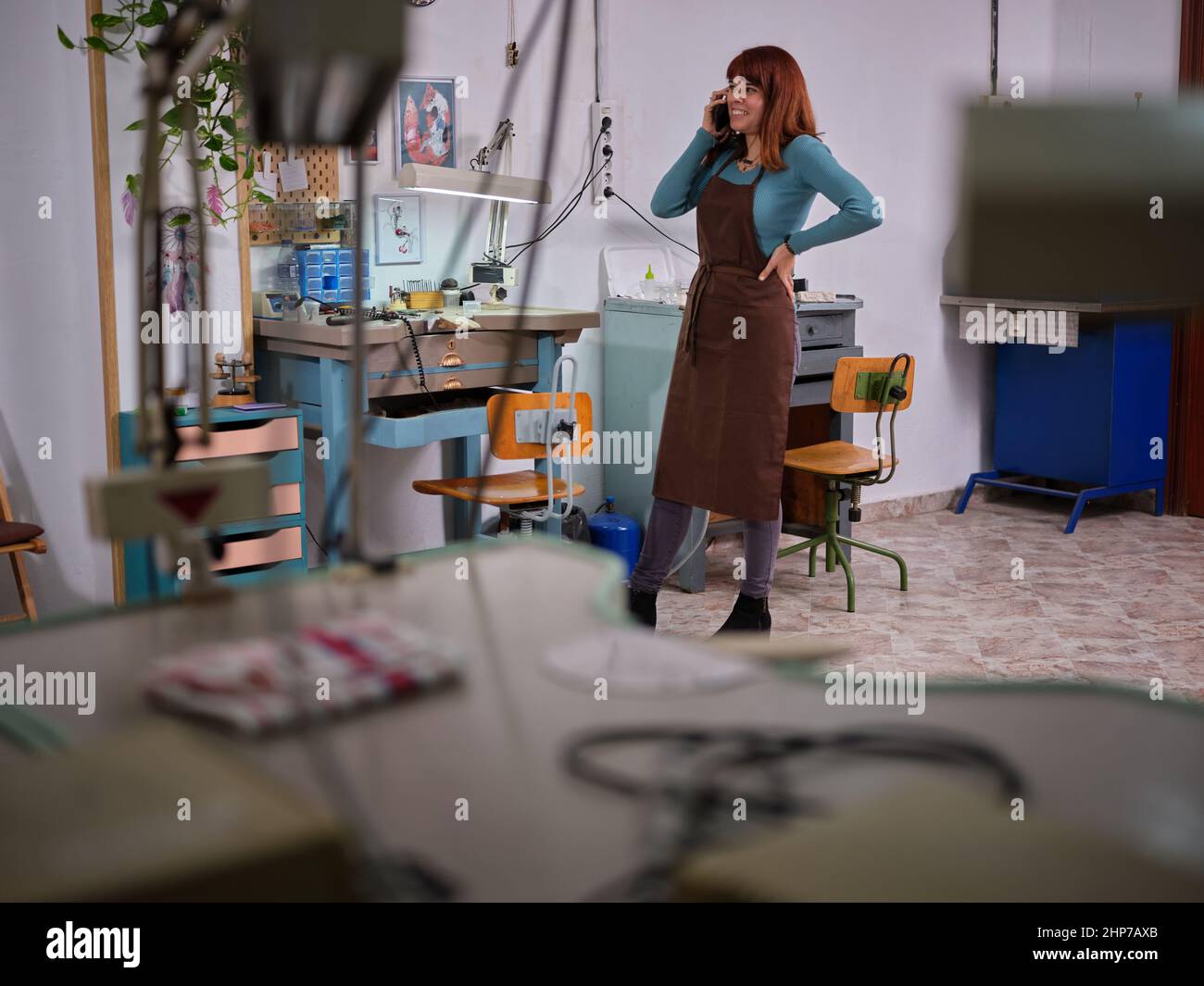 Eine Arbeiterin mit Schürze, die in ihrer Schmuckwerkstatt steht, während sie mit ihrem Mobiltelefon spricht Stockfoto
