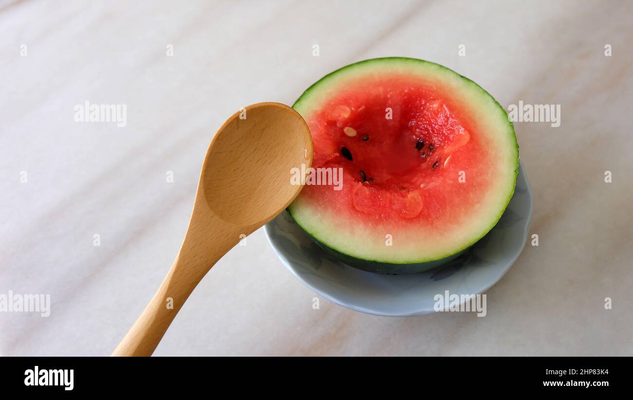 Kleine Wassermelone in zwei Hälften geschnitten, mit einem kleinen Loch in der Mitte und einem Holzlöffel daneben. Auf einer Marmoroberfläche. Stockfoto