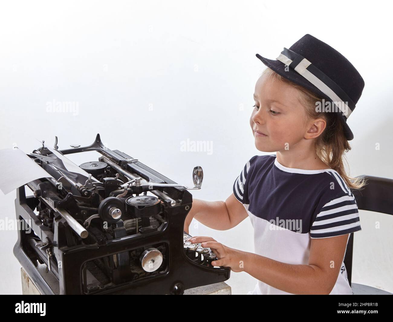 Hübsches kleines Mädchen im Hut, das auf antiker Schreibmaschine auf weißem Hintergrund tippt. Bildungskonzept.fcool Stockfoto