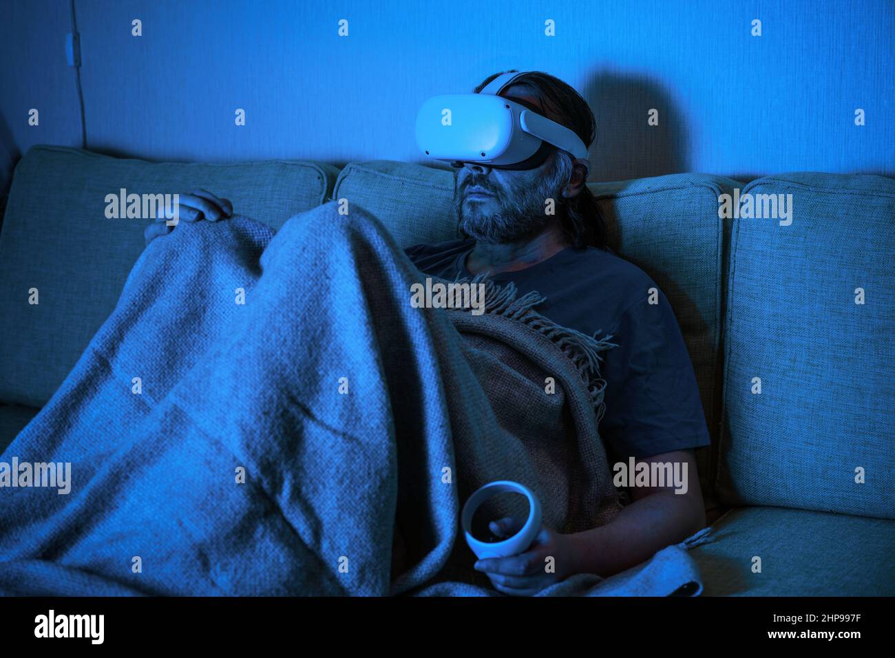 Der Mann, der auf dem Sofa sitzt, nutzt nachts ein Virtual-Reality-Headset, eine traurige Person, die allein zu Hause in einer VR-Brille schaut. Konzept der Einsamkeit, Fernsehen, entspannen Stockfoto