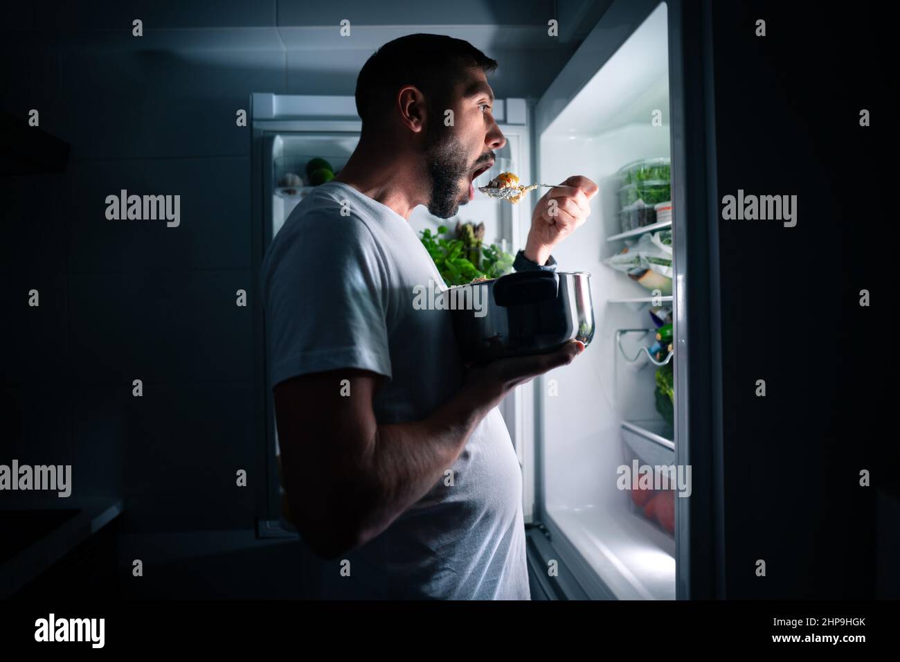 Hungriger Mann, der nachts aus dem offenen Kühlschrank Essen isst. Mann, der Mitternachtssnack aus dem Kühlschrank nahm Stockfoto
