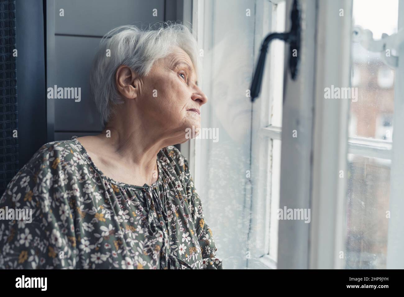 Seitenaufnahme einer älteren europäischen Großmutter, die während des Tages der Großeltern ganz allein war und sanft aus dem Fenster schaute. Hochwertige Fotos Stockfoto