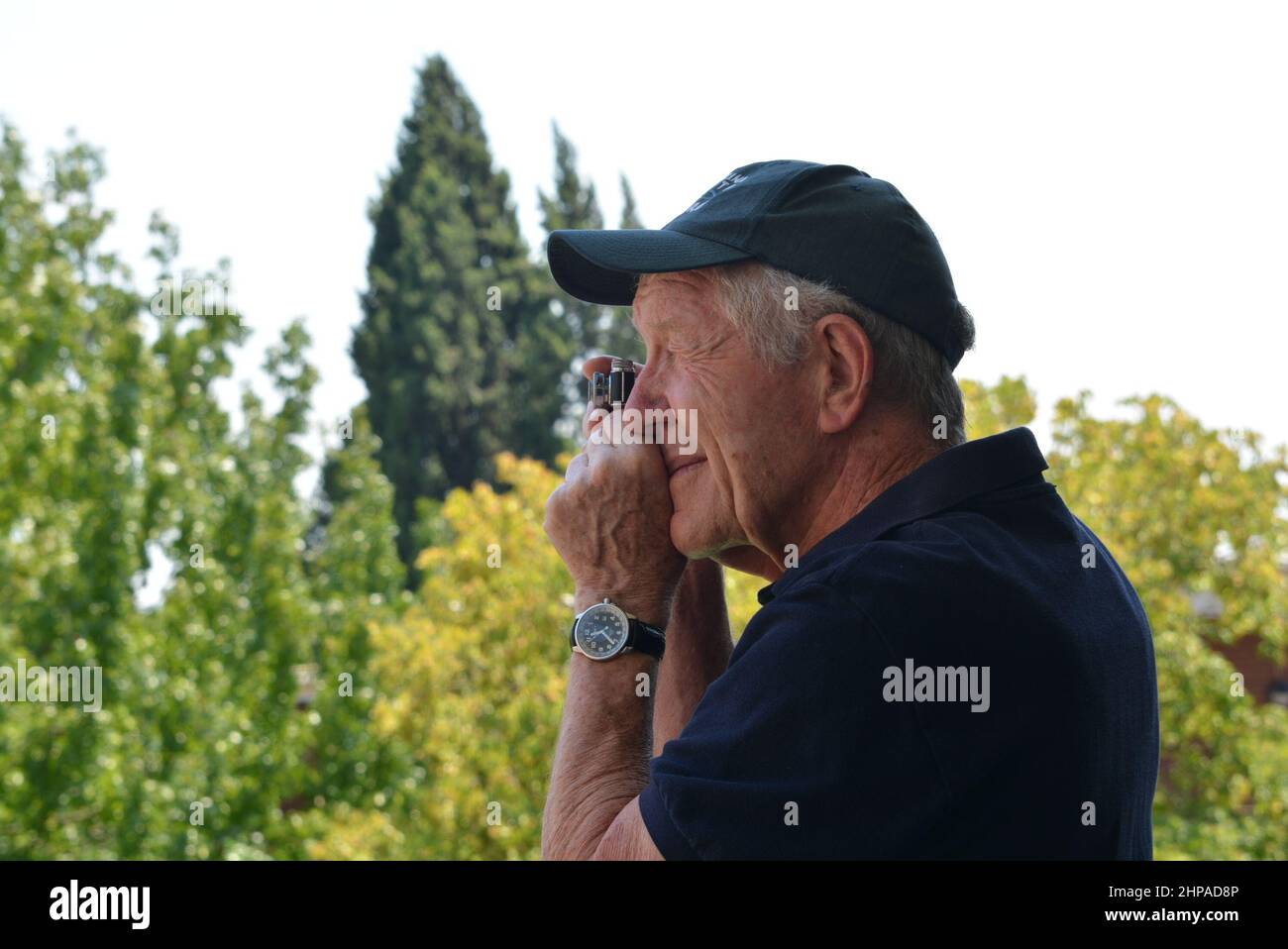 Älterer Fotograf mit einer Miniatur-Filmkamera auf Augenhöhe. Blaues Hemd mit blauer Mütze auf einem weichen Hintergrund von grünen Bäumen. Stockfoto
