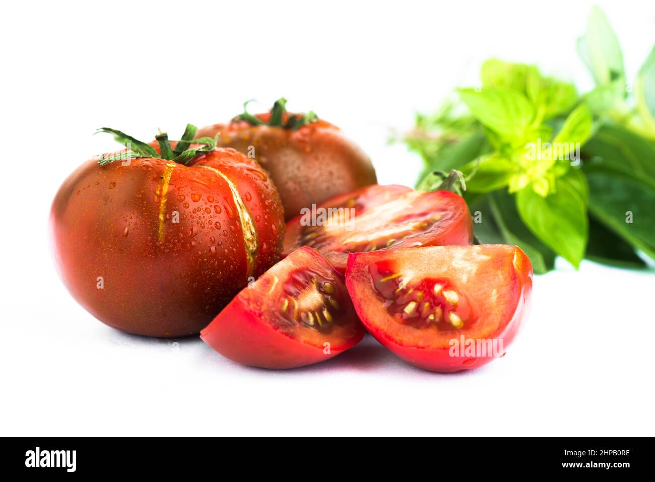 Ganze und halbierte dunkelrote taufige Tomate mit frischem grünen Basilikumblättern, isoliert auf weißem Hintergrund. Stockfoto