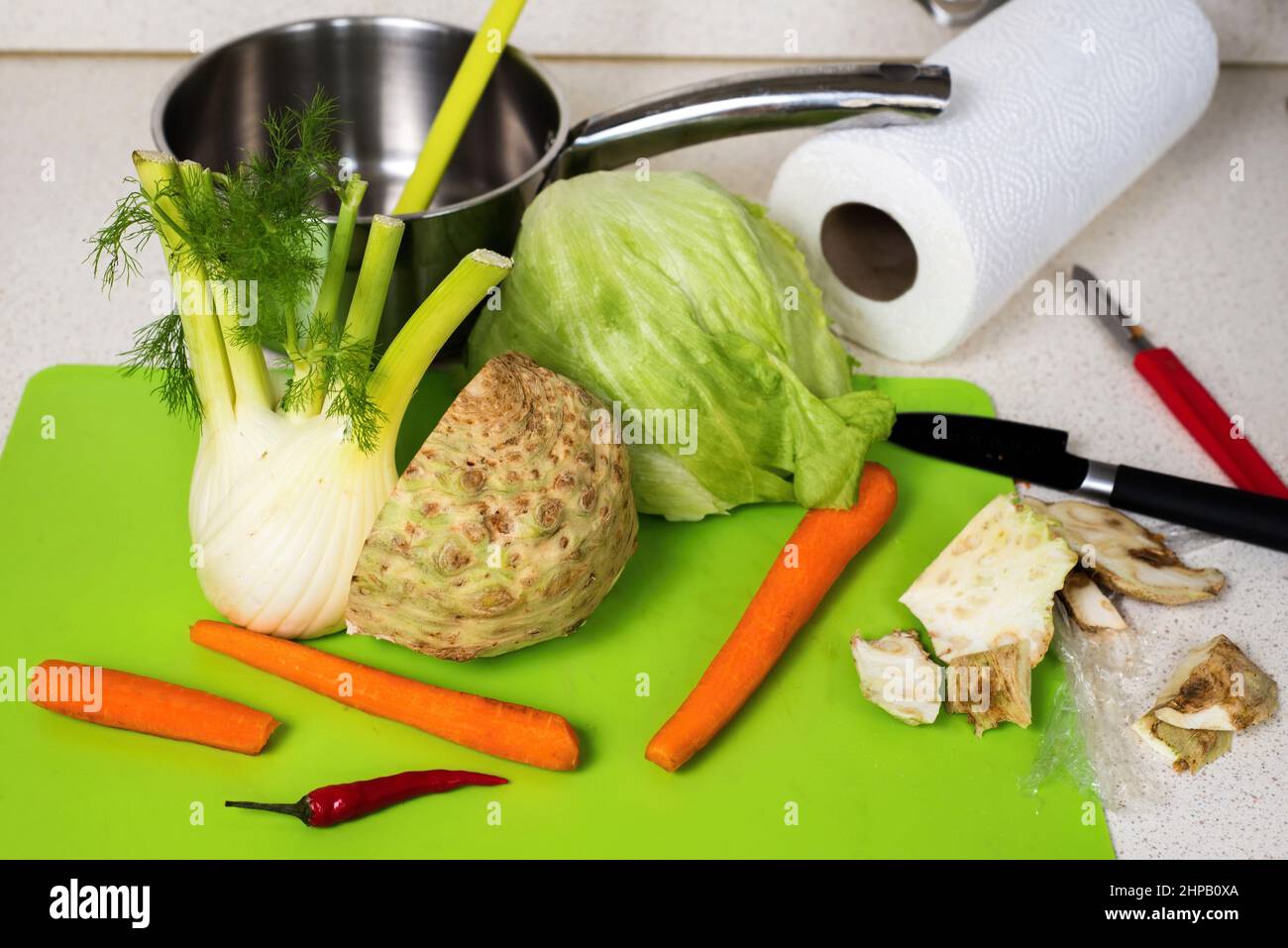 Rohes Gemüse (Sellerie, Karotten, Salat und Zichorie) auf grünem Küchentafel mit Messer, Papierhandtuchrolle und abschälender Sellerie. Suppe wird zubereitet. Stockfoto