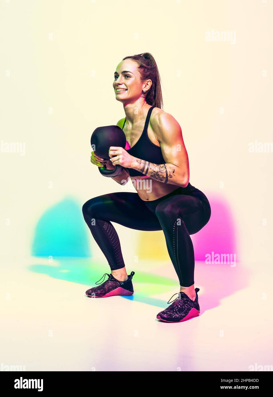 Sportliche Frau macht tiefe Kniebeugen Übung mit Kettlebell. Foto von Frau in schwarzer Sportkleidung auf weißem Hintergrund mit Effekt von rgb-Farben Schatten. Str Stockfoto