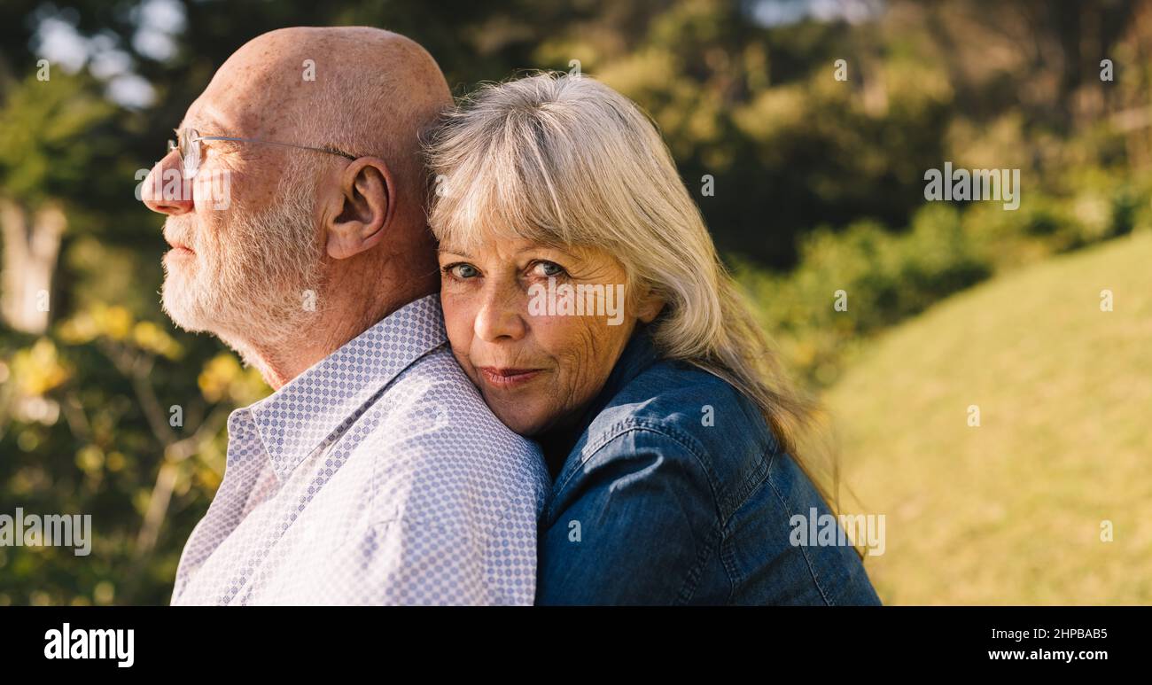 Schönes älteres Paar, das sich in einem Park umarmt. Romantische ältere Frau, die hinter ihrem Mann steht und auf die Kamera schaut. Reifes Paar Stockfoto