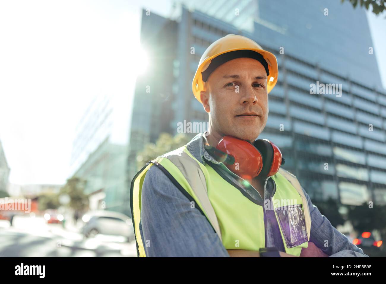 Selbstbewusster Arbeiter, der die Kamera ansah, während er mit gekreuzten Armen in der Stadt stand. Bauarbeiter mit mittlerem Erwachsenenalter, der vor einem Hochhaus steht Stockfoto