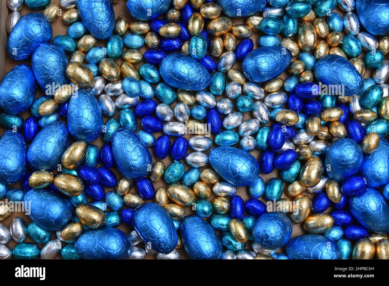 Kleine, in hellen und dunkelblauen, silbernen und goldenen Folien verpackte Ostereier mit Schokolade, mit großen blauen Eiern vor einem hellen Holzhintergrund. Stockfoto