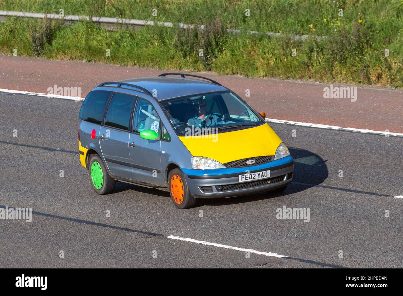 2002 mehrfarbige, maßgeschneiderte, personalisierte Ford Galaxy LX TDI 6-Gang-Schaltgetriebe, Autoverpackung, Vinyl-Grafiken auf farbigem Car Wrapping mit gelber Motorhaube und unterschiedlich farbigen Rädern; Fahrt auf der Autobahn M6, Großbritannien Stockfoto