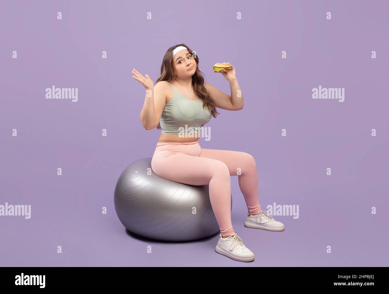 Unsicher, traurige und große europäische Millennial-Dame in Sportbekleidung, die zweifelt, ob es Burger auf Fitnessball sitzt Stockfoto