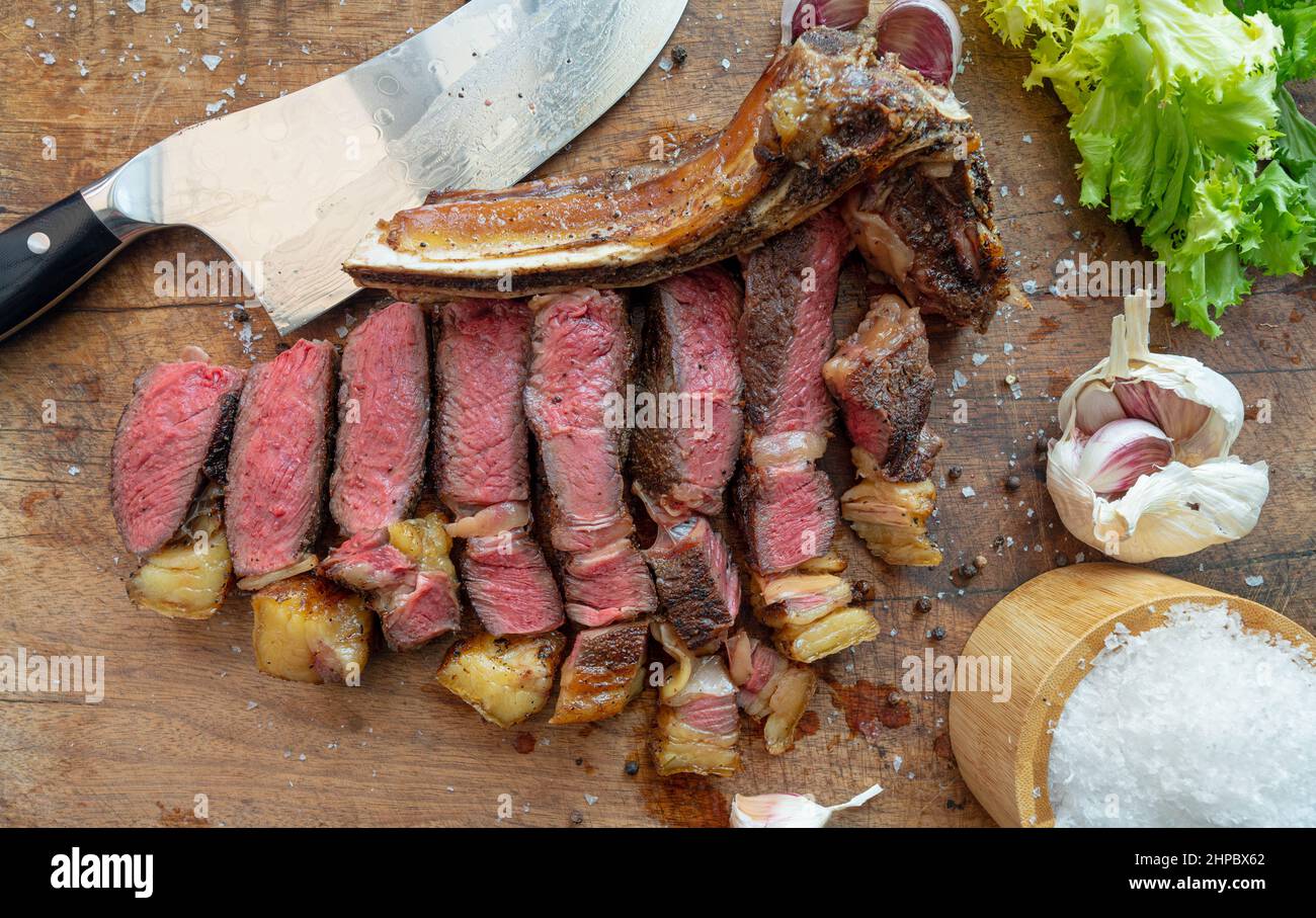 In Scheiben geschnittenes Sous-Vide gekochtes, mittelseltenes, trocken gereiftetes rubia-Gallega-Rindersteak mit Messer, Meersalzflocken, Knoblauch, Pfeffer und Salat auf einem Holzbrett Stockfoto