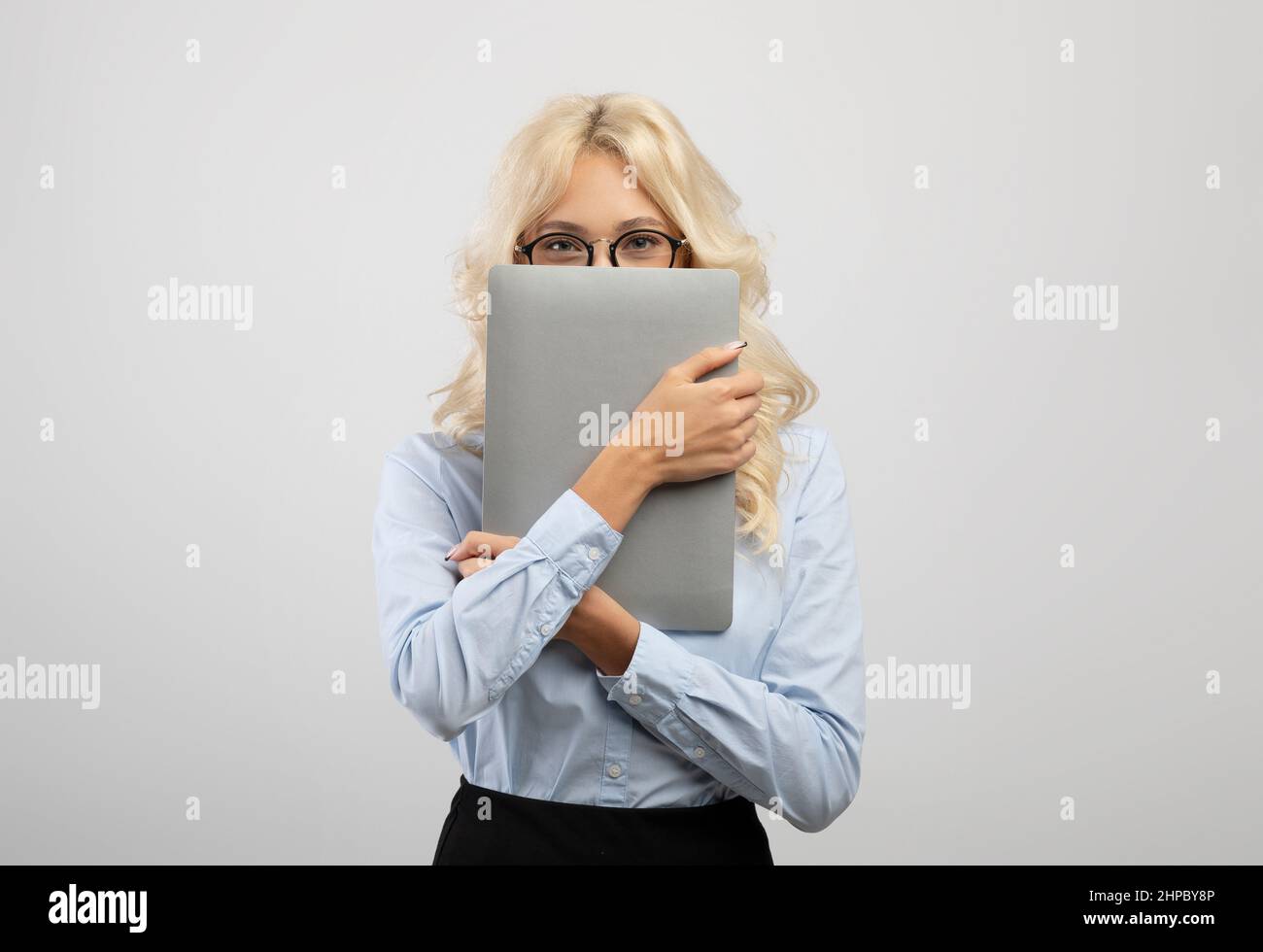 Erstaunte junge Geschäftsfrau, die das Gesicht mit einem Laptop bedeckt, formelle Kleidung trägt und auf hellem Studiohintergrund posiert Stockfoto