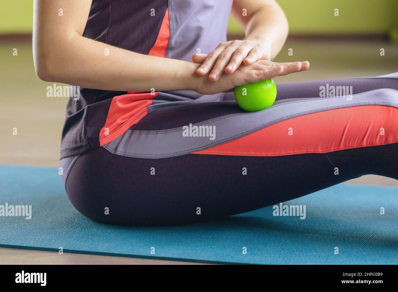 Frau in Sportbekleidung, führt mit einem Massageball myofasziale Entspannung der Oberschenkelmuskulatur durch, sitzt auf einer Matte im Raum, Nahaufnahme Stockfoto